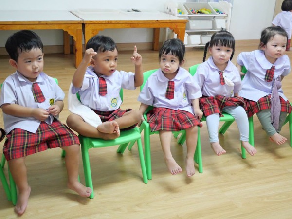 Five children in a row on green chairs, one with a hand up