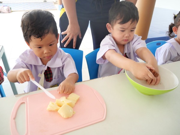 Two children cutting fruit on a board, a teacher standing behind them