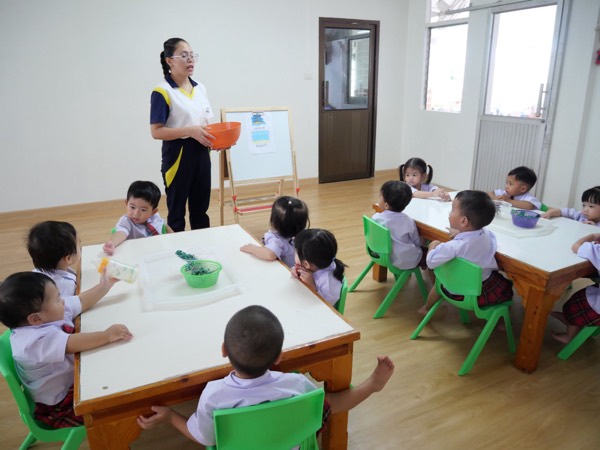 A bright classroom, children at two tables, the teacher standing at the front with a bowl