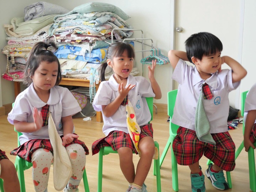 椅子に座って歌に合わせて体を動かす三人の子ども。後ろにはたたんだ寝具