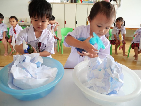 Two children leaning over two basins, spraying colour onto crumpled white cloth