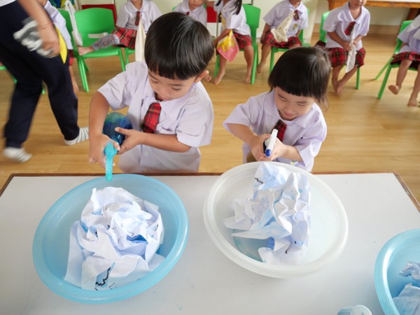 Two children spraying colour onto cloth in a basin, friends waiting behind them