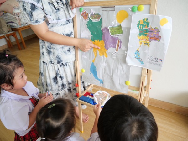 A teacher pointing at a painted map of Thailand on an easel while the children watch